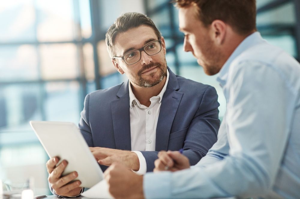Two professional men in business attire discuss work, with one holding a tablet, in a bright, modern office setting.