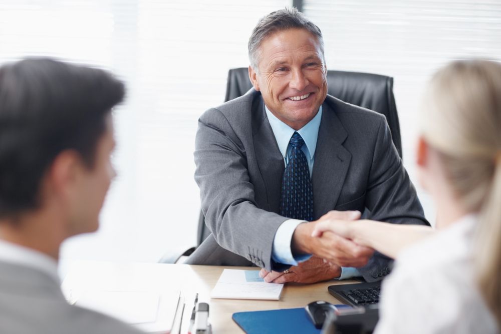 Man in suit shaking hands with a woman at a desk; another person is also present.