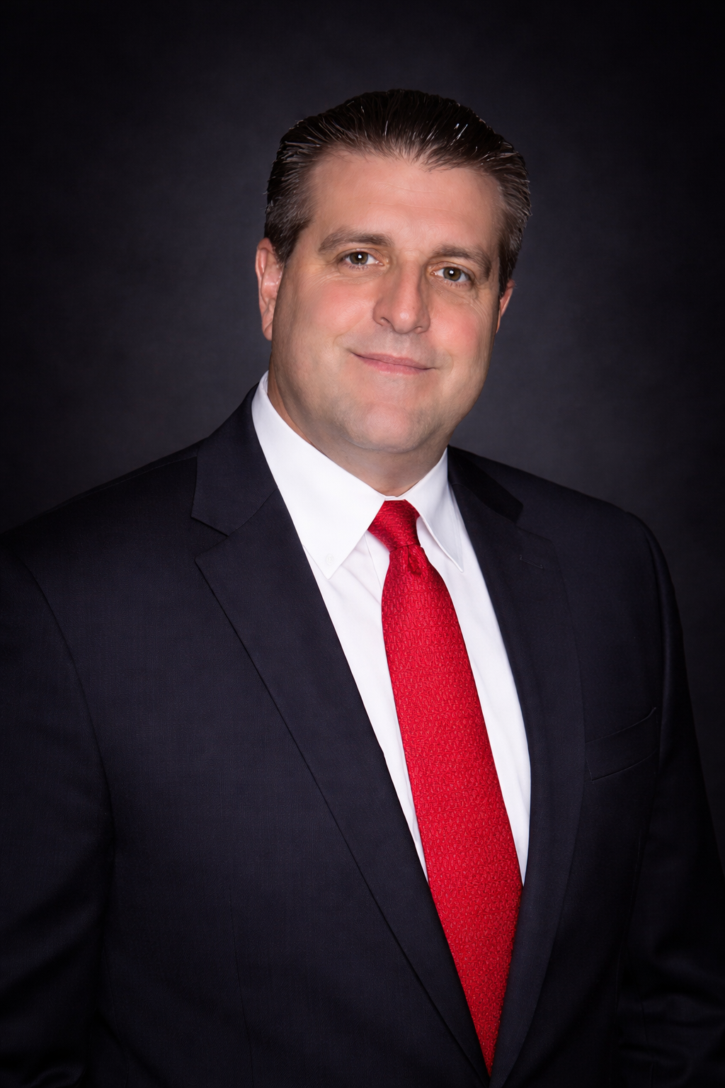 A headshot of a person in a dark suit, white shirt, and red tie, posing against a dark, neutral background.