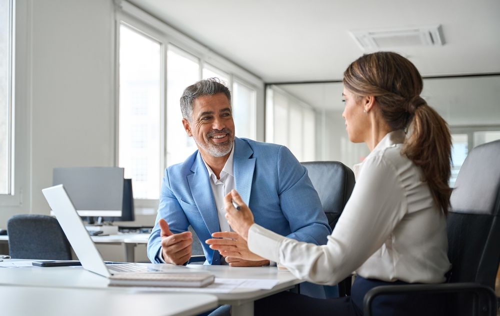 Man and woman in office, discussing, gesturing. Smiling, laptop on desk. Bright room with windows.