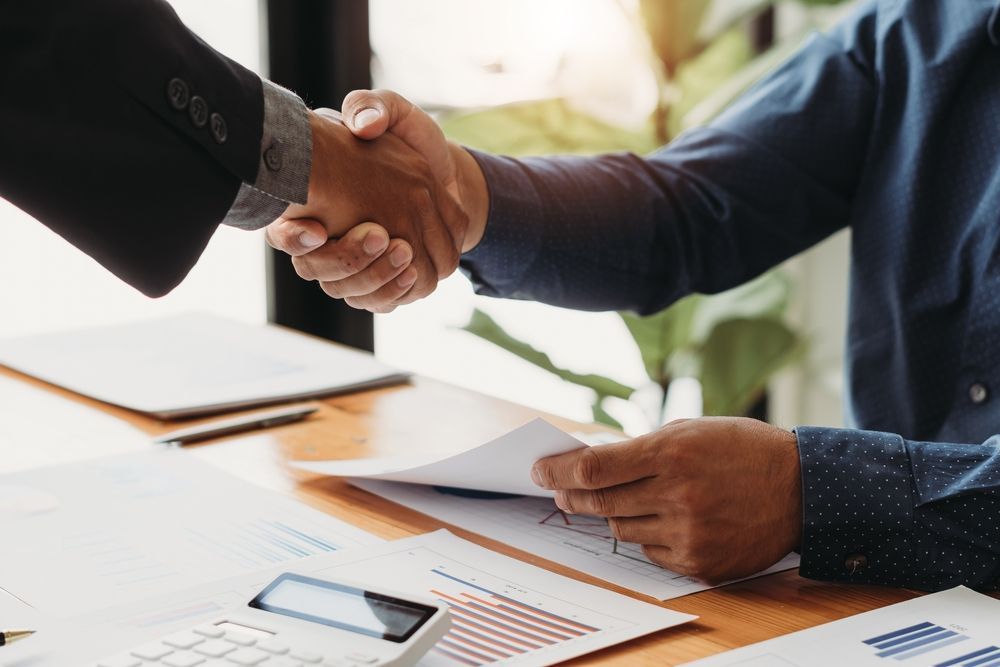Two people shaking hands over a table with papers, a calculator, and a tablet.