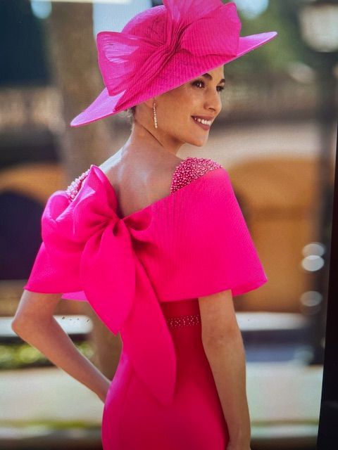 Woman in fuchsia dress with large bow and matching hat, smiling, outdoors.