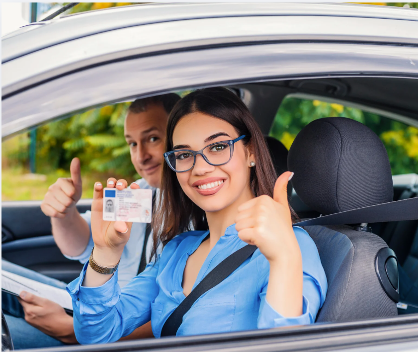 A smiling driver in a blue shirt holds up a driver's license, giving a thumbs up alongside an instructor in a car.