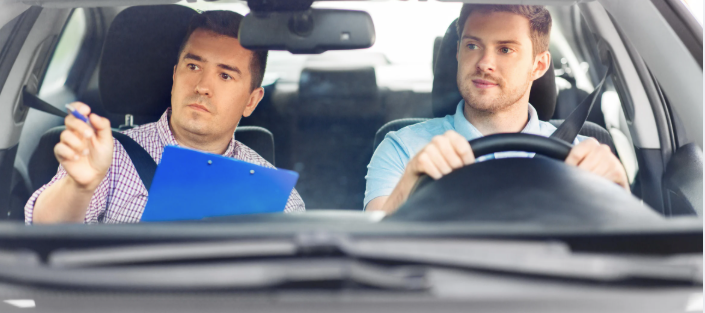 A driving instructor holding a blue clipboard gives directions to a person driving a car.