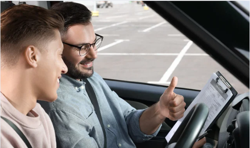 An instructor gives a thumbs up to a student while holding a clipboard inside a car.
