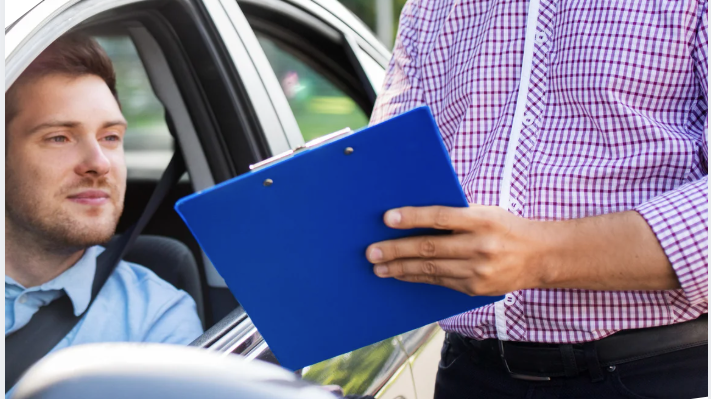 A person in a checkered shirt holds a blue clipboard while talking to a driver sitting inside a car.