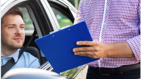 A person in a checkered shirt holds a blue clipboard while talking to a driver sitting inside a car.