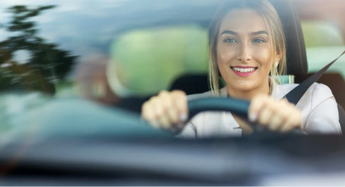 A smiling person driving a car, hands on the steering wheel, wearing a seatbelt.
