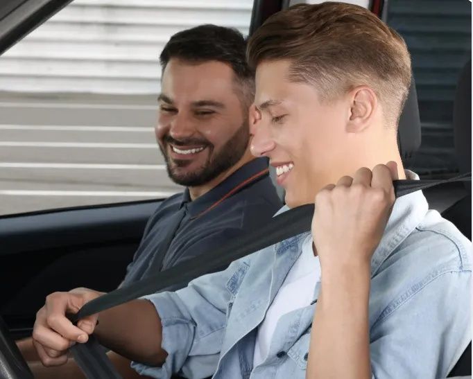 A person seated in a car fastens their seatbelt while a smiling passenger looks on from the seat behind.