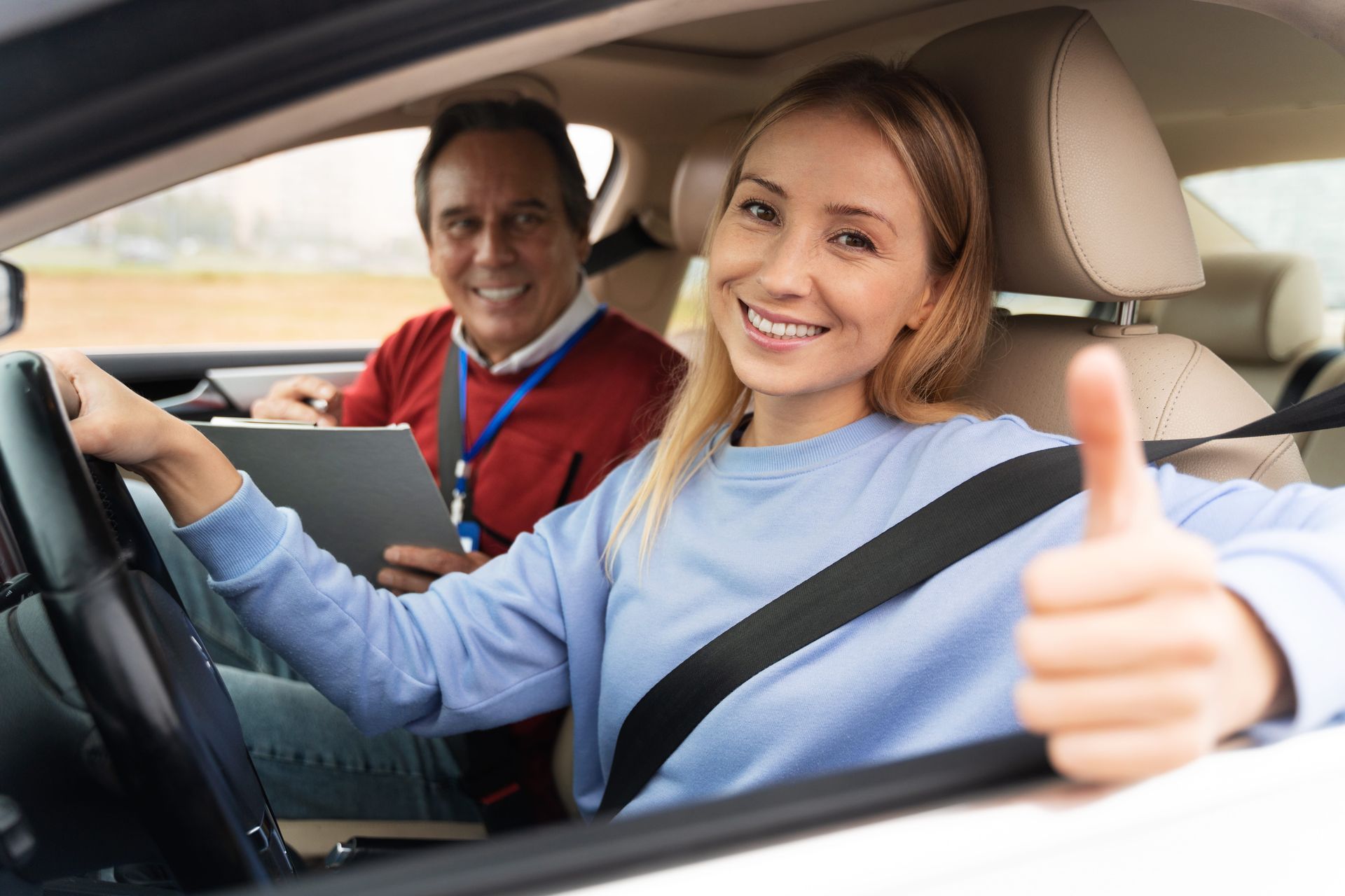 A driving instructor points at the road while a student steers a car during a lesson, viewed from the backseat.