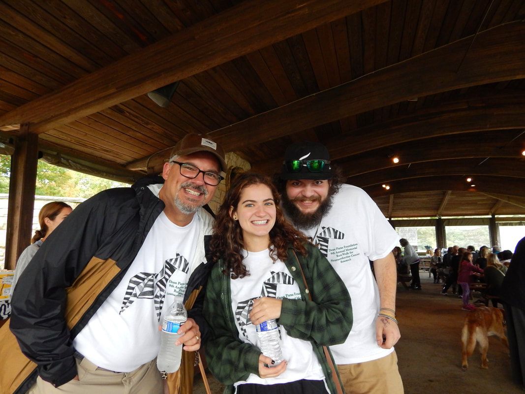 Three people smiling under a wooden shelter, wearing matching t-shirts. Two men flank a woman.