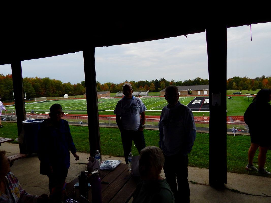 People under a shelter, overlooking a field. Cloudy sky. Some are standing, others seated at a picnic table.