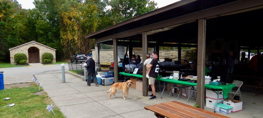 People gathered under a pavilion with tables, dog, and a small building in the background.