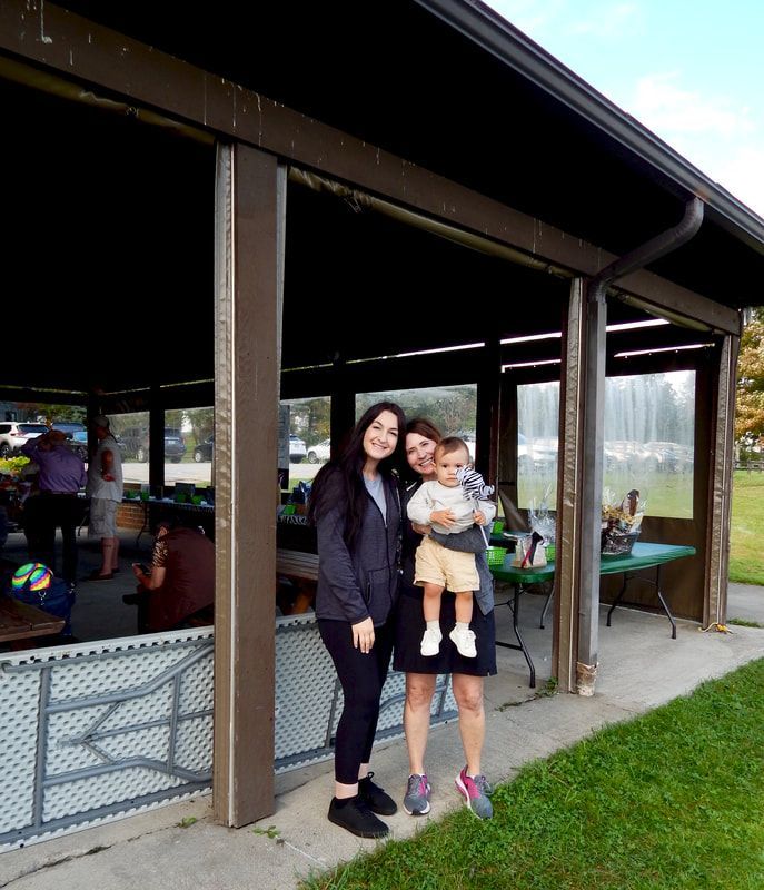 Three people smiling at a park pavilion. One holds a toddler. Green grass and fountain in background.