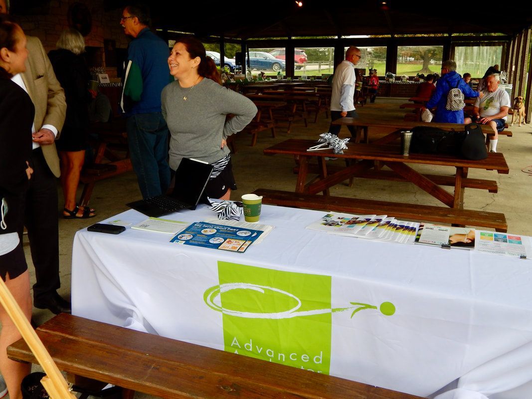 People at a table with a white tablecloth, logo, and brochures in an outdoor pavilion.