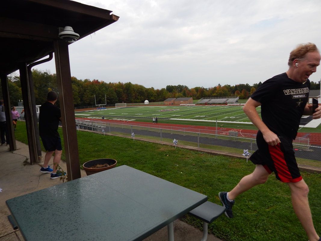 Man running outside, near a green field and stadium. Wearing black shirt and red shorts.