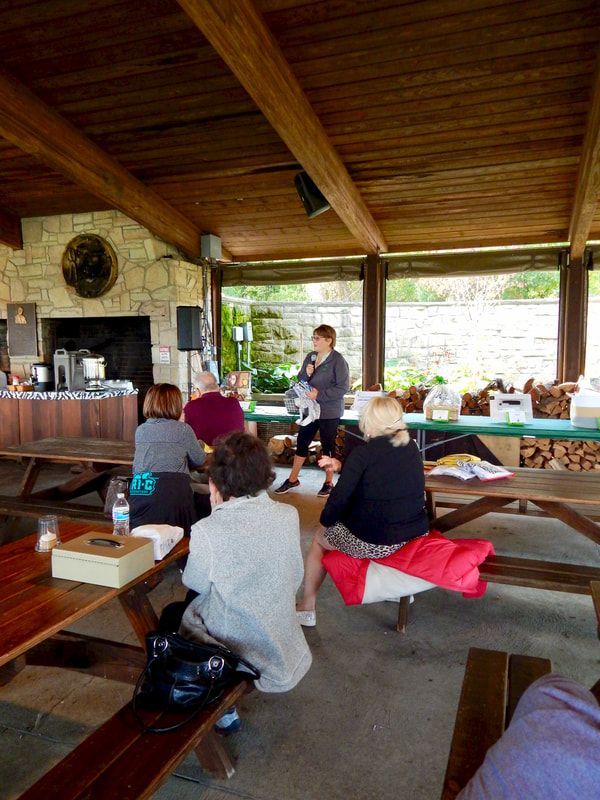 People seated at picnic tables listen to a woman speaking outdoors under a covered structure.