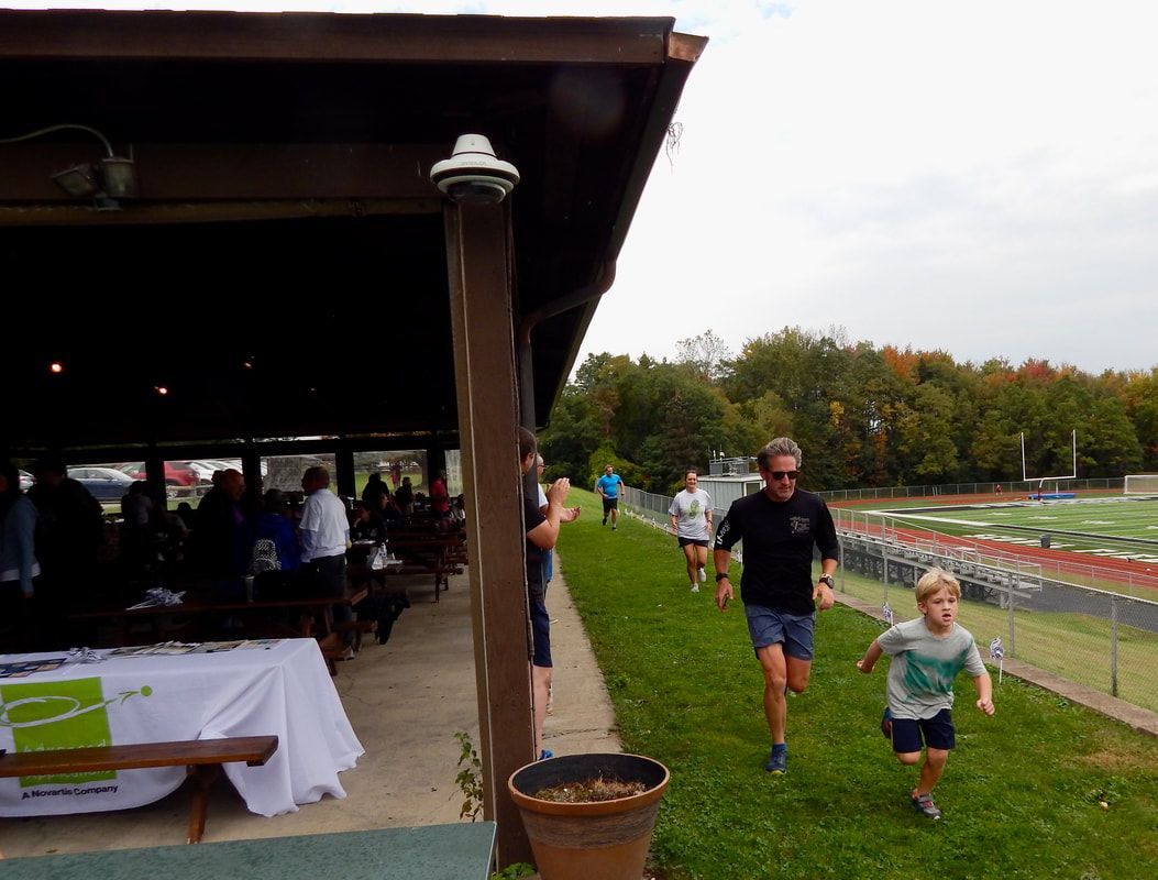 People running on grass, near a covered pavilion. A child runs in front of a man. Trees in background.