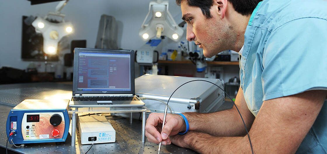 Dean Paras in a lab coat looks at a monitor, holding a small tool. He's surrounded by lab equipment.