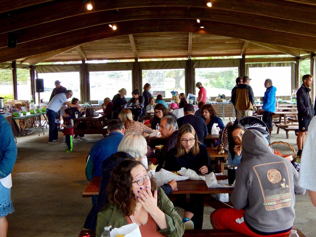 People gather at picnic tables under a covered pavilion for an outdoor event.