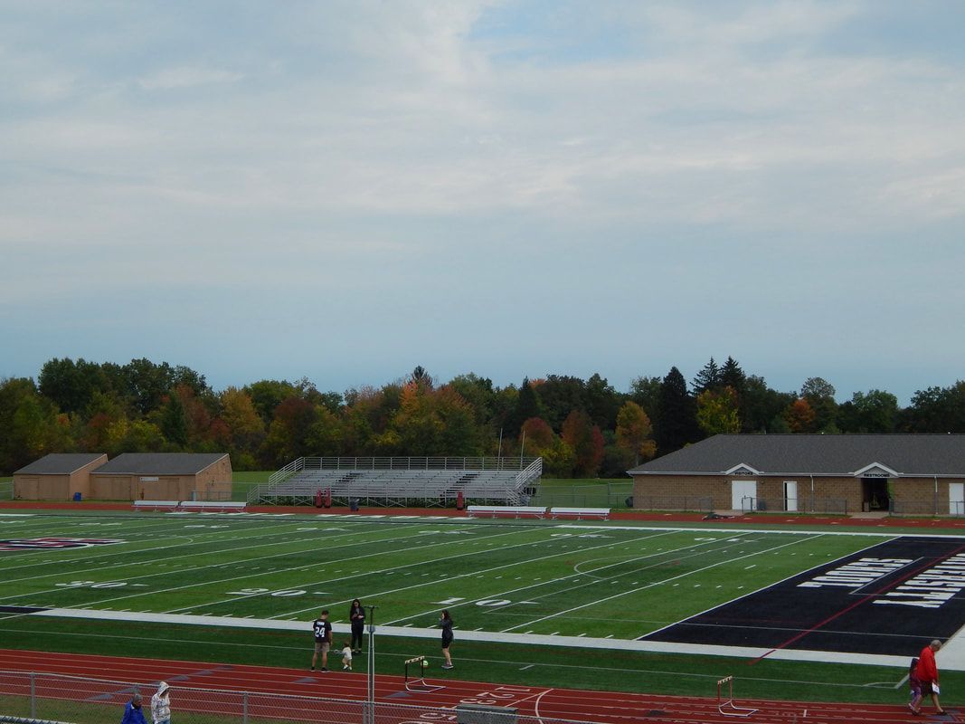 Football field with bleachers, buildings, and track under a cloudy sky.