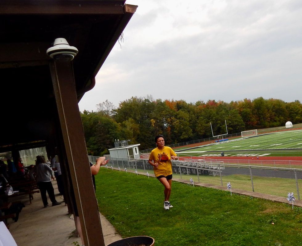 A runner in a yellow shirt sprints on grass near a track and football field. Trees and a cloudy sky in background.