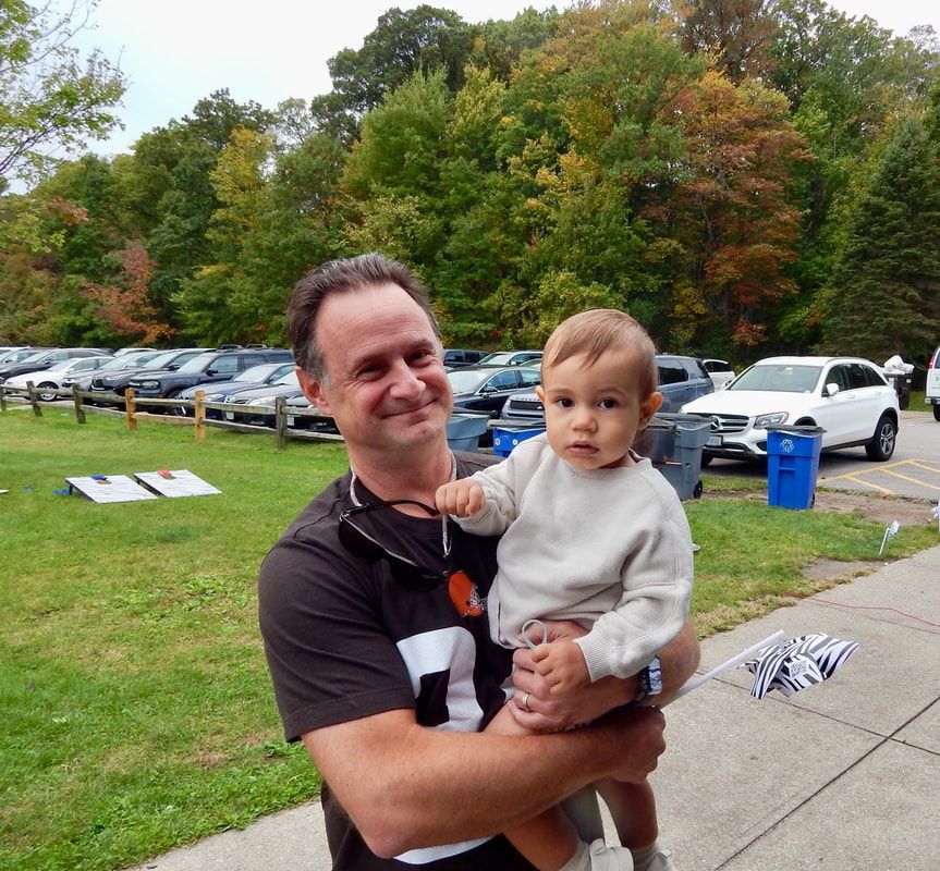 Man holding a baby outdoors; trees and cars in the background. They are both looking at the camera.