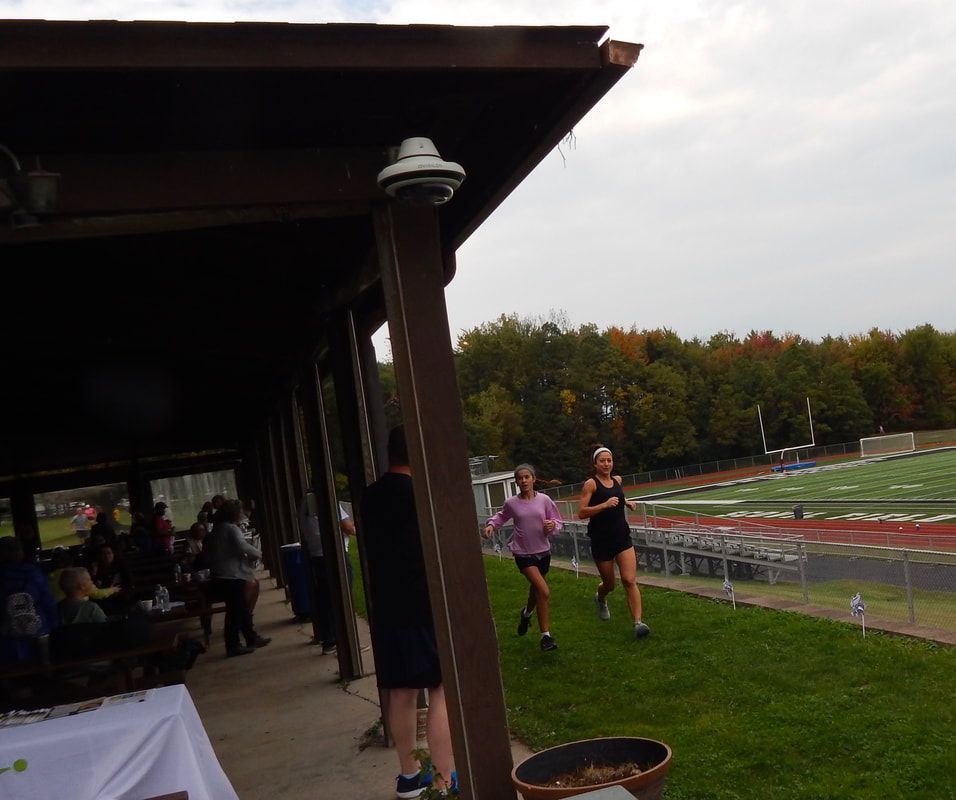 Two runners on grass near a covered structure and a football field. Fall foliage in background.