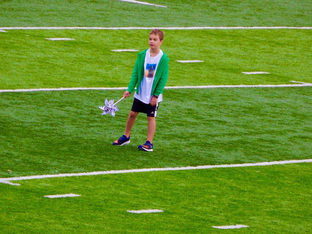 Boy on a football field holding a pinwheel; he wears green sweater, black shorts, and sneakers.