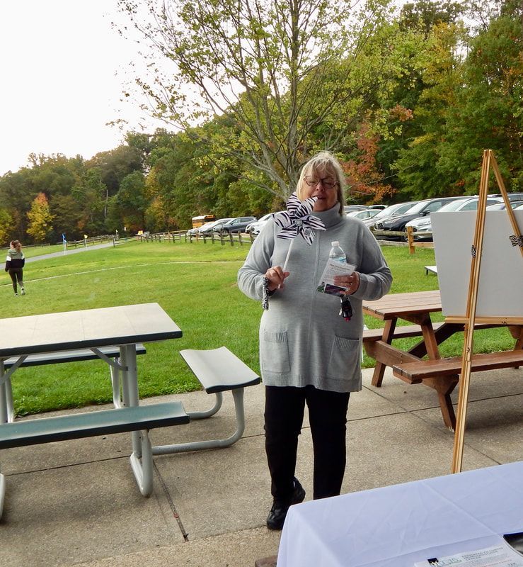 Woman standing outdoors holding items near a picnic table and easel with a green grassy area and trees in the background.