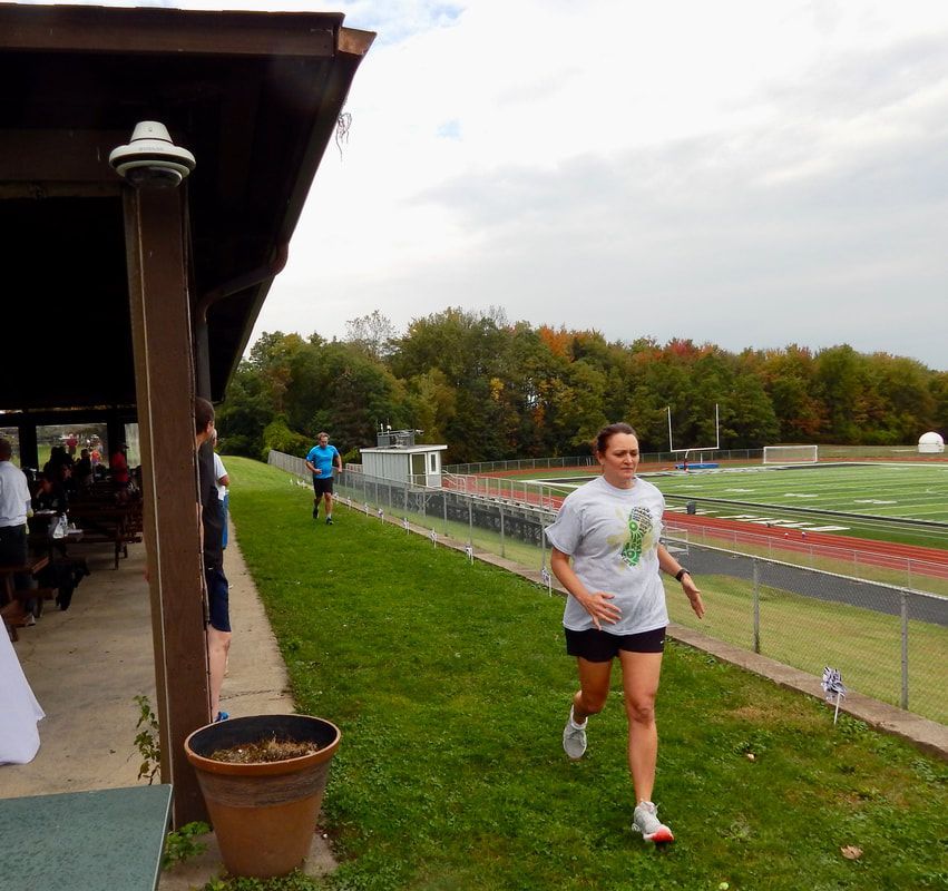 Woman in grey shirt runs on grass by a pavilion and a sports field.