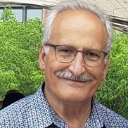 Man with glasses and mustache smiling in front of green foliage.