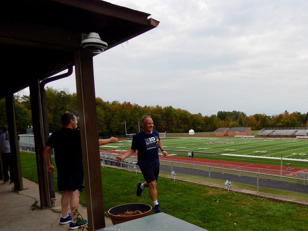 Two men running near a football field under a cloudy sky. One smiling.