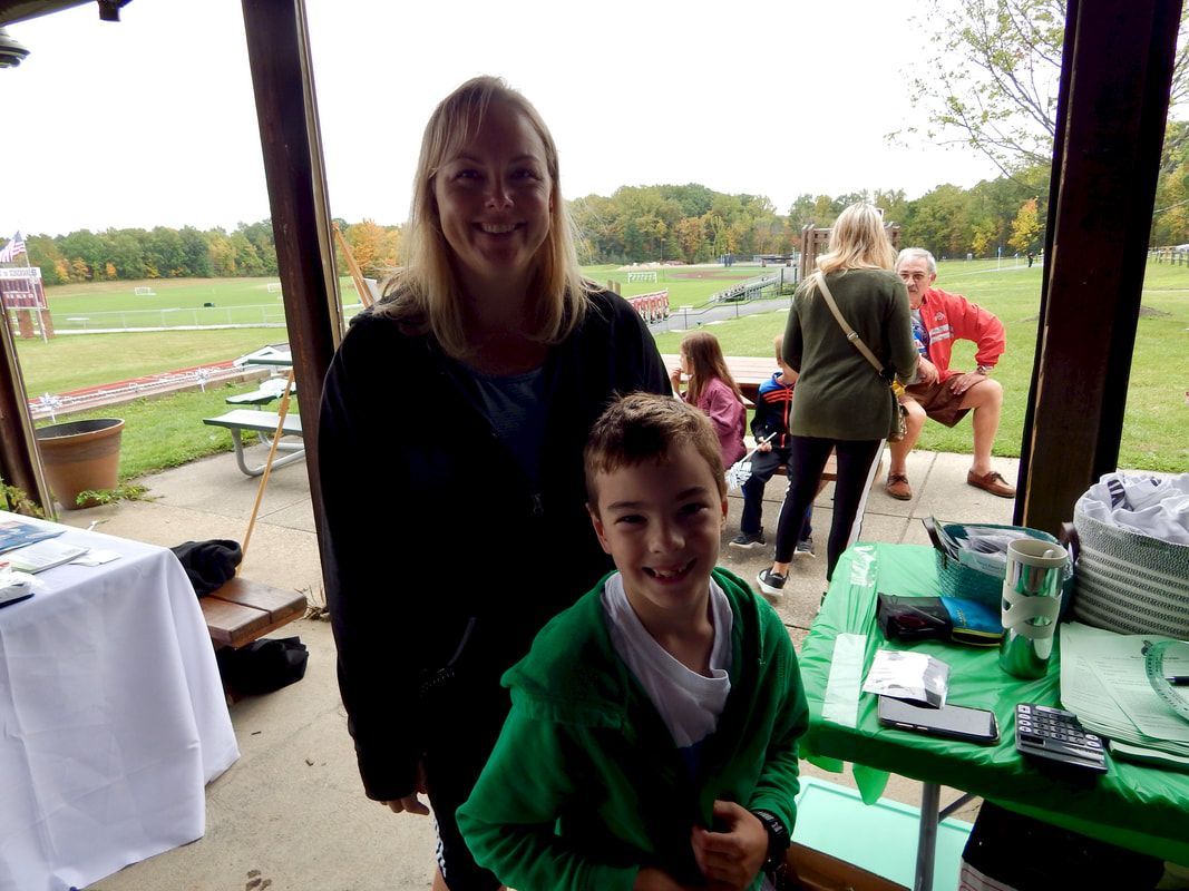 Woman and child smiling at the camera under a covered outdoor area with other people in the background.