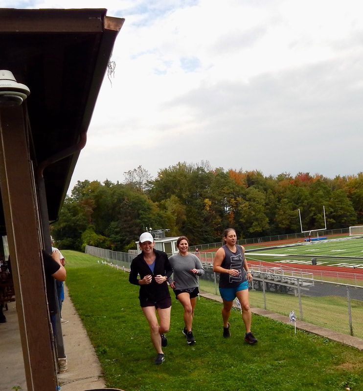 Three people running on grass next to a track under an overcast sky.
