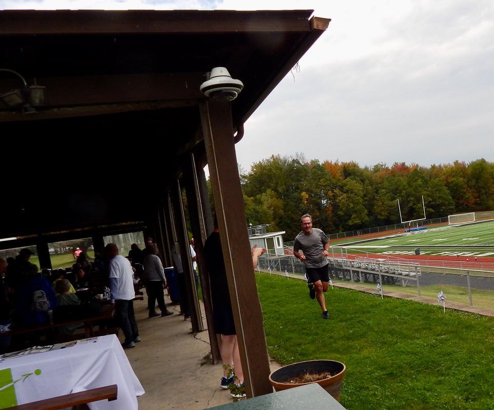 Man running on grass near a covered pavilion with people and a track and field in the background.