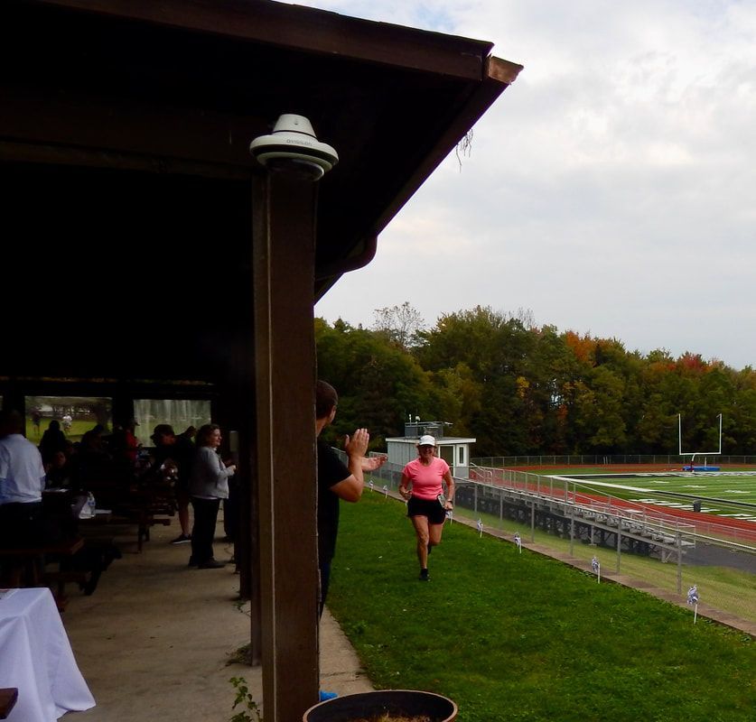Runner in pink shirt approaching a football field, people clapping from a covered area, trees in the background.