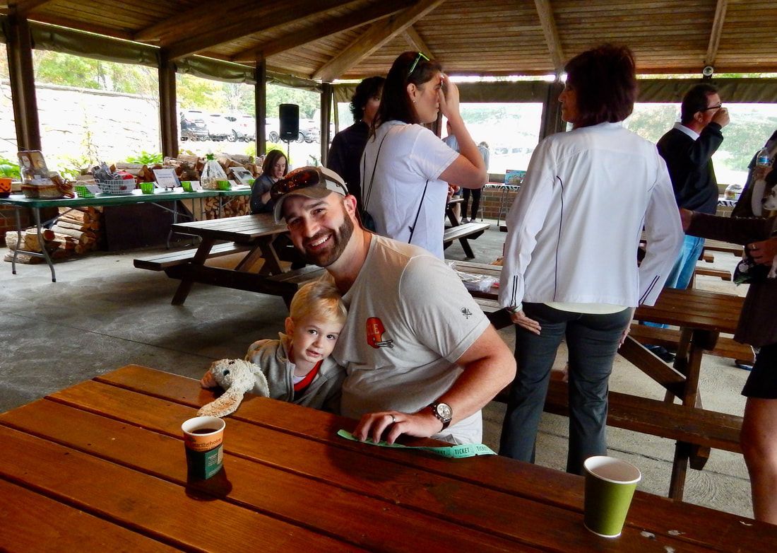 Man and child at a picnic table, smiling. Other people stand nearby in an outdoor pavilion.