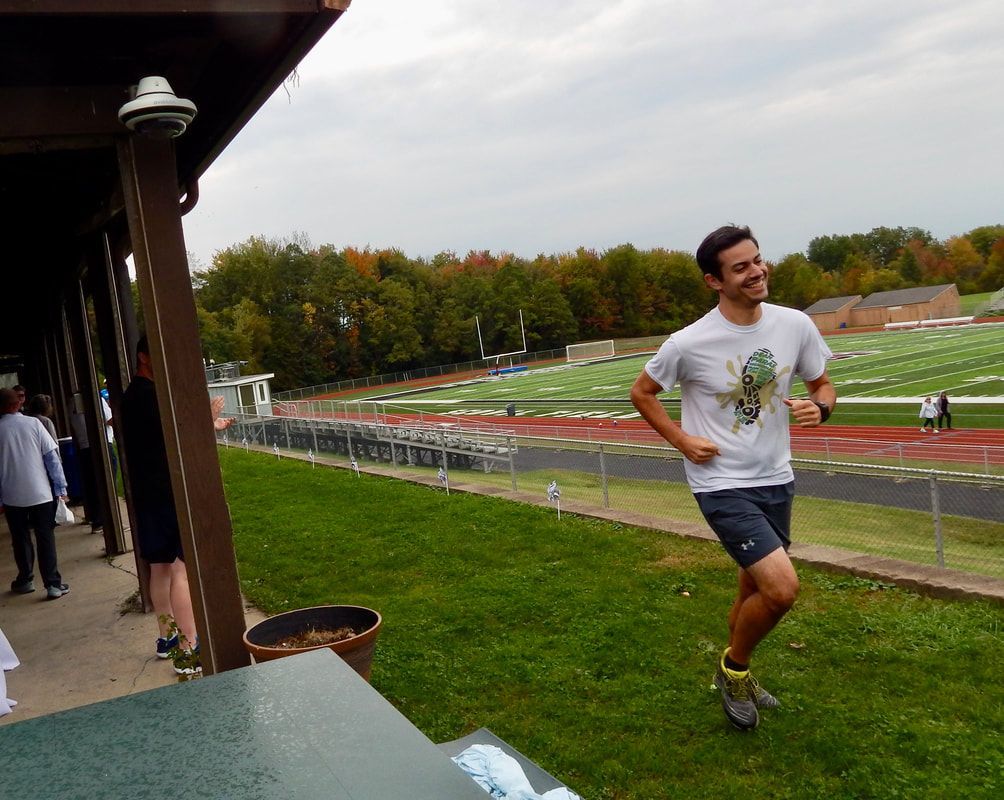 Man running on grass near a track, smiling. Wearing gray shirt, dark shorts, green and gray shoes. Cloudy day.
