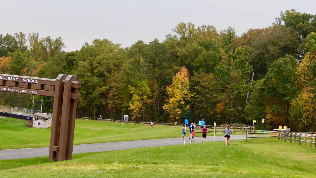 Runners on a green path through a park, flanked by fall foliage and a wooden structure on a cloudy day.