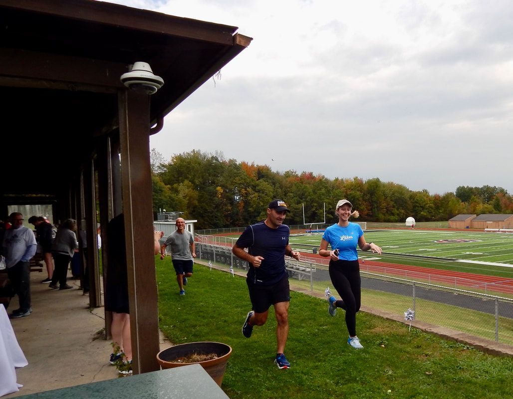 People running on a grassy area, near a building and track. Overcast sky.