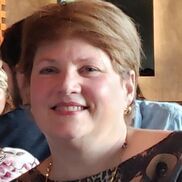 Woman with short blonde hair smiles, wearing patterned top, and seated indoors.