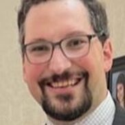 Man with glasses smiling, wearing a button-down shirt and suit jacket, indoors.