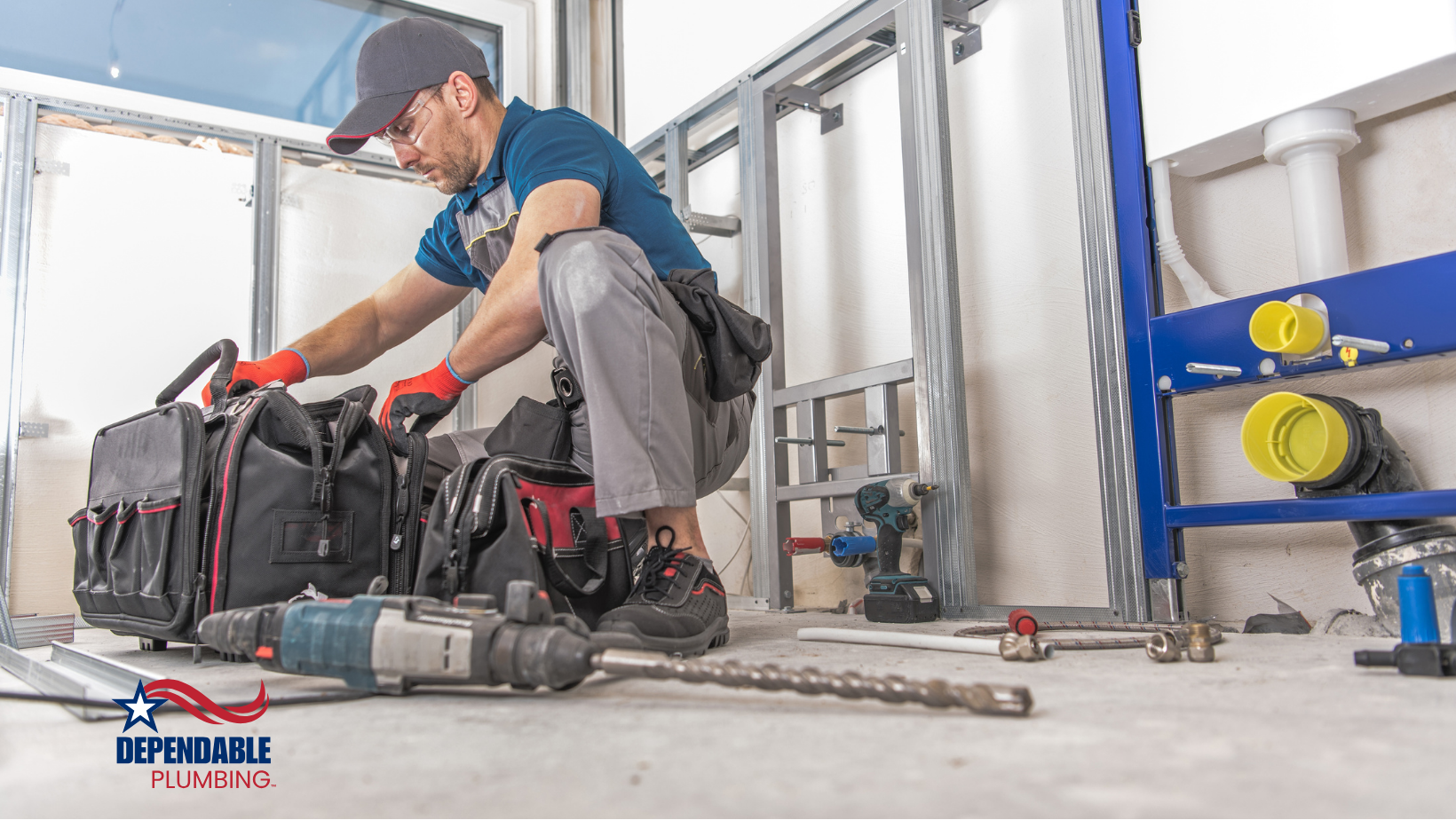 A plumber in a blue shirt kneels near his tool bag, with tools and plumbing in a construction site.