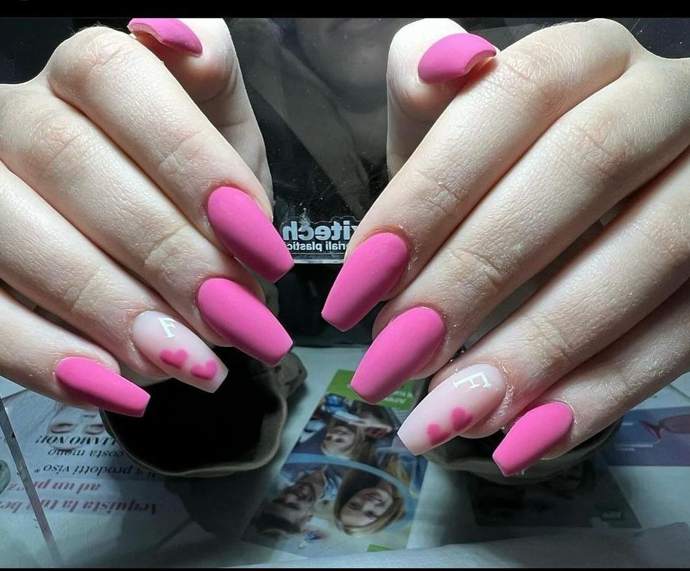 Mani che mostrano una manicure con unghie rosa opache e due unghie decorate con piccoli cuoricini rosa.