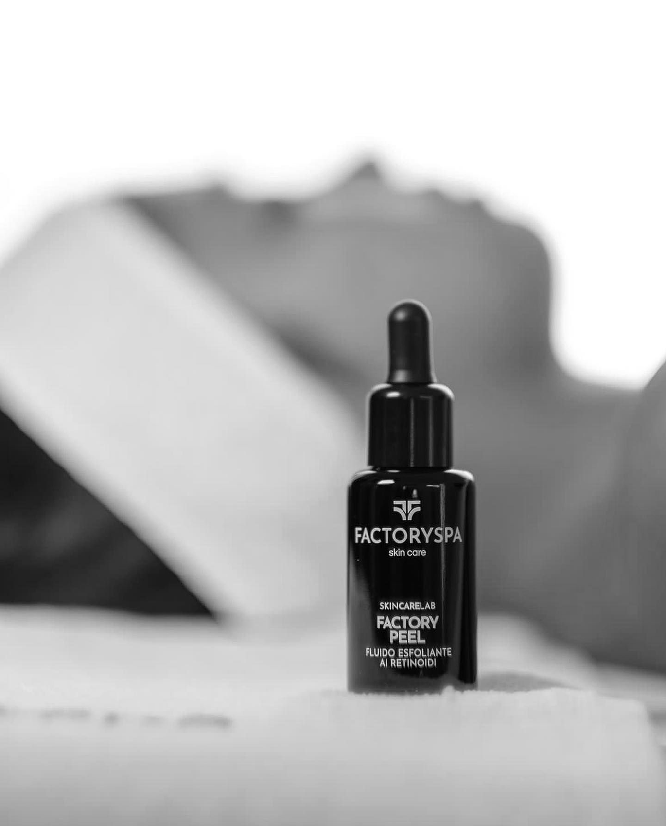 Primo piano in bianco e nero di una boccetta contagocce con l'etichetta "Factory Spa" appoggiata su un asciugamano bianco, con una persona sullo sfondo.