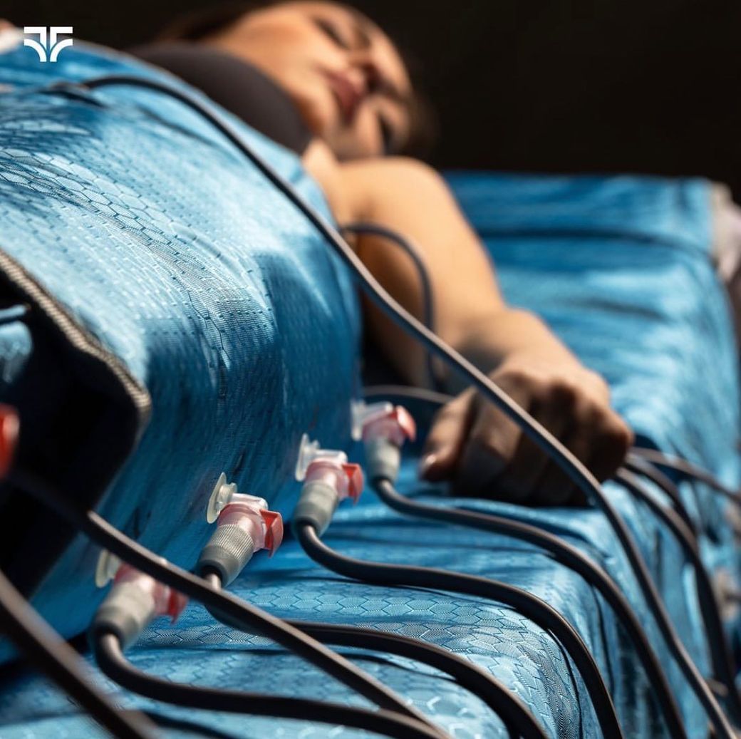 Una persona è sdraiata su un lettino indossando una tuta blu per pressoterapia, collegata tramite tubi neri a un apparecchio.