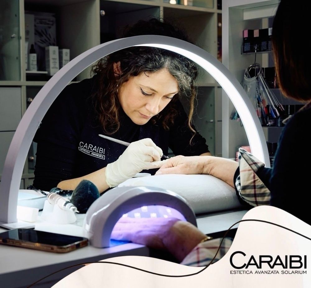 Un'estetista esegue una manicure a una scrivania illuminata da una lampada ad arco, con la mano della cliente all'interno di una lampada per unghie.