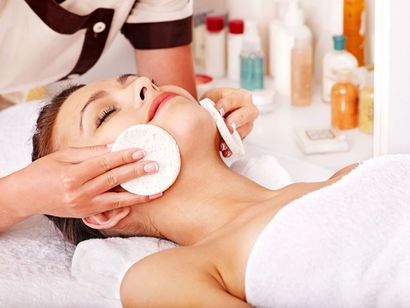 In un ambiente spa, un'estetista professionista deterge il viso di una cliente con dischetti di cotone, con diversi prodotti per la cura della pelle sullo sfondo.