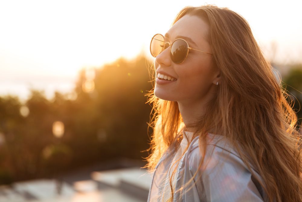 Woman with sunglasses smiles, looking off to the side as the sun sets behind her — Eyepower In Tahmoor, NSW